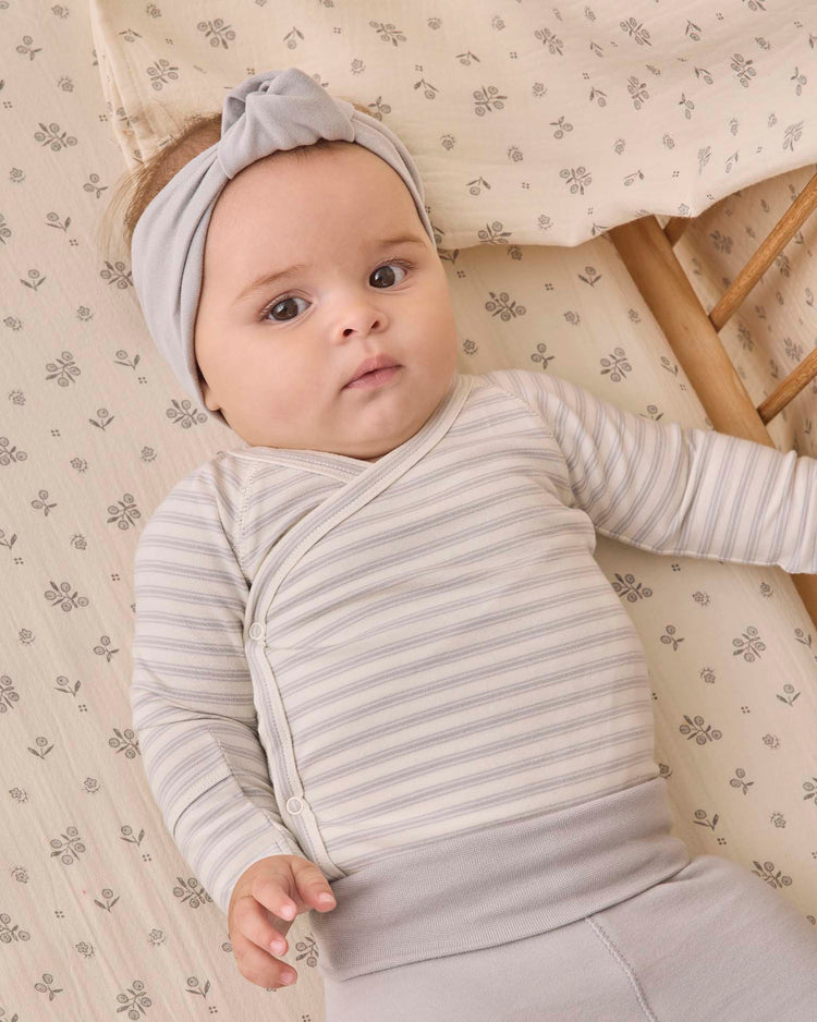 A baby wearing the quincy mae side snap bodysuit + pant set in cloud stripe, along with a gray headband, lies on a patterned crib sheet and gazes slightly upward with a calm expression.