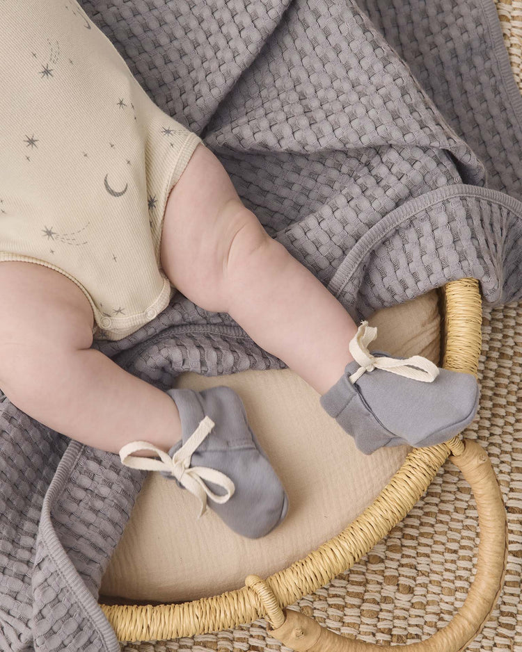A baby wearing a beige onesie decorated with moon and star patterns and quincy mae baby booties in slate lies on a textured gray organic cotton blanket inside a woven basket on a woven mat.