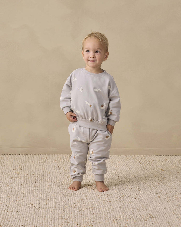 A toddler stands barefoot on a textured rug, smiling and wearing the quincy mae relaxed sweatshirt in sky friends—a light gray matching set with a celestial pattern, crafted from soft organic cotton—against a beige background.
