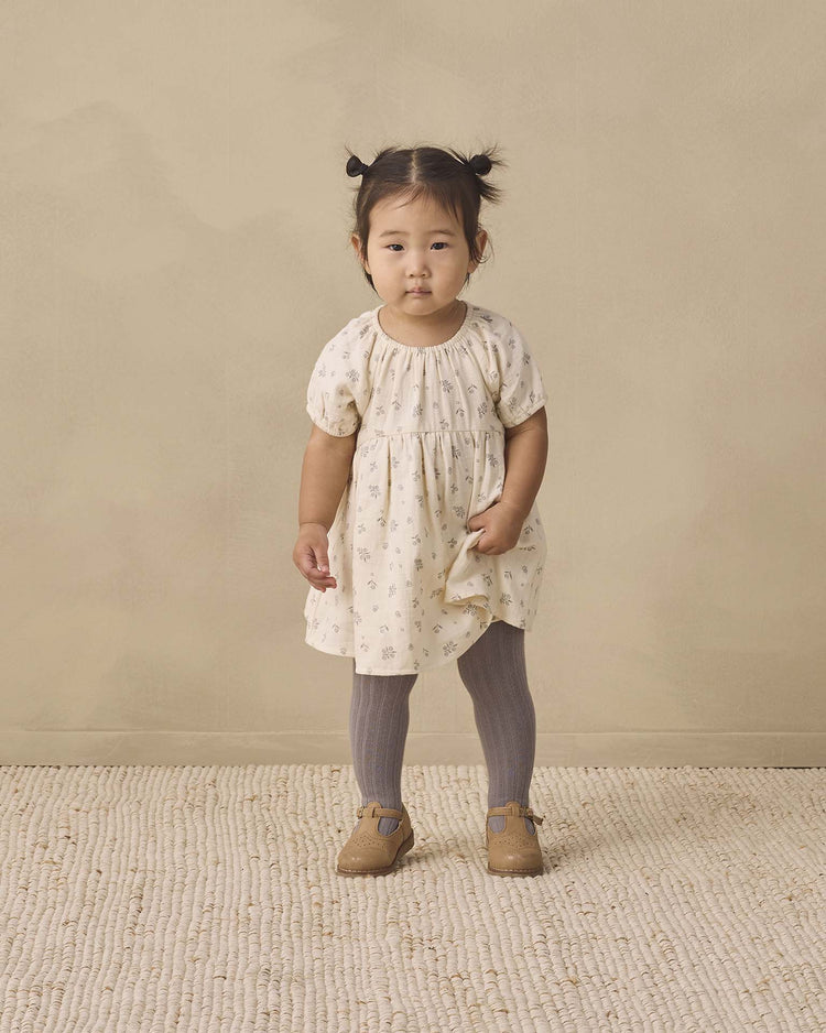 A young child with pigtails stands on a textured rug against a beige background, wearing the quincy mae bella dress in winter berry, paired with gray tights and brown shoes.