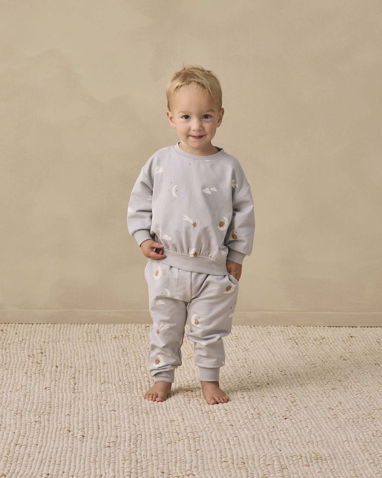 A toddler with light hair stands barefoot on a textured rug, wearing the quincy mae relaxed sweatshirt in sky friends—crafted from organic cotton and adorned with a whimsical moons and clouds print—paired with matching pants, set against a plain beige background.