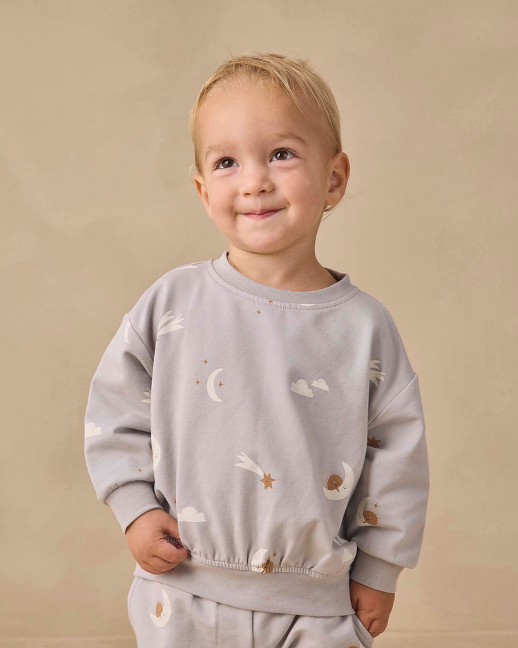 A young child with short blond hair smiles, wearing the quincy mae relaxed sweatshirt in sky friends—a light gray organic cotton piece adorned with a charming moons, stars, and clouds print—standing against a plain beige background.