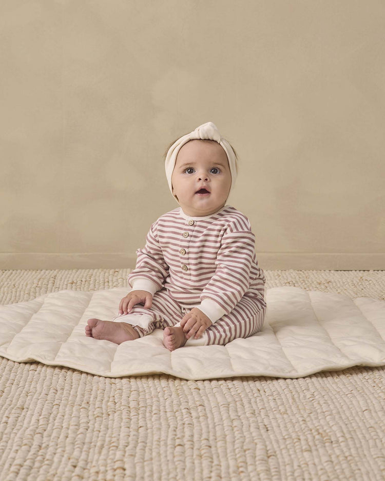 A baby wearing the quincy mae relaxed jumpsuit in mulberry stripe, paired with a matching headband, sits on a cream quilted mat atop a textured beige rug, gazing slightly upward with a curious expression.