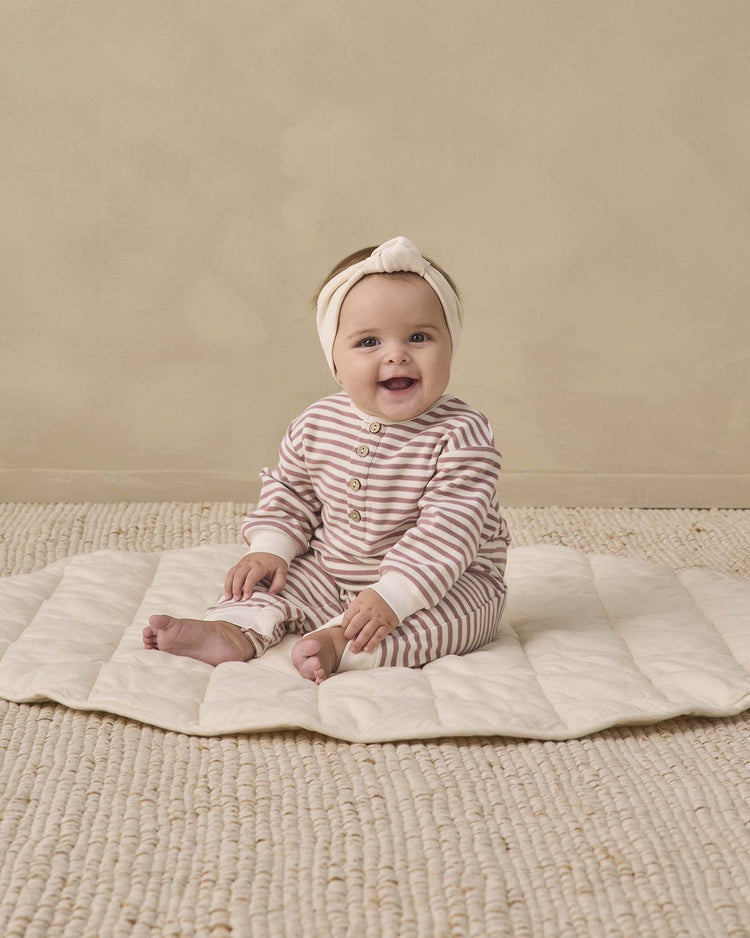 A smiling baby in the quincy mae relaxed jumpsuit in mulberry stripe, paired with a matching headband, sits on a cream quilted mat atop a textured beige rug, set against a plain beige wall.