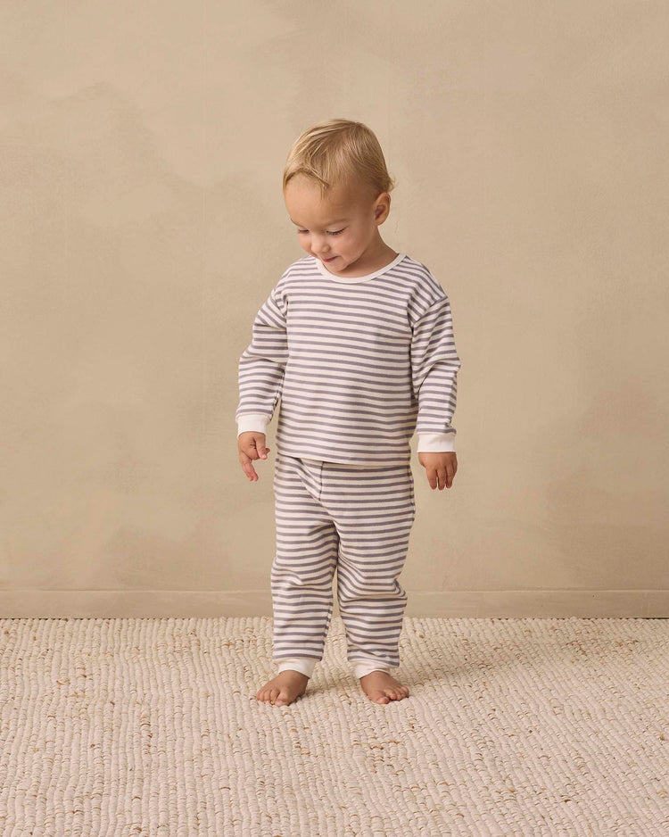 A toddler stands barefoot on a textured rug, dressed in the quincy mae cozy set in slate stripe—matching striped pajamas with drawstring pants. The child gazes downward with a slight smile against a plain beige background.