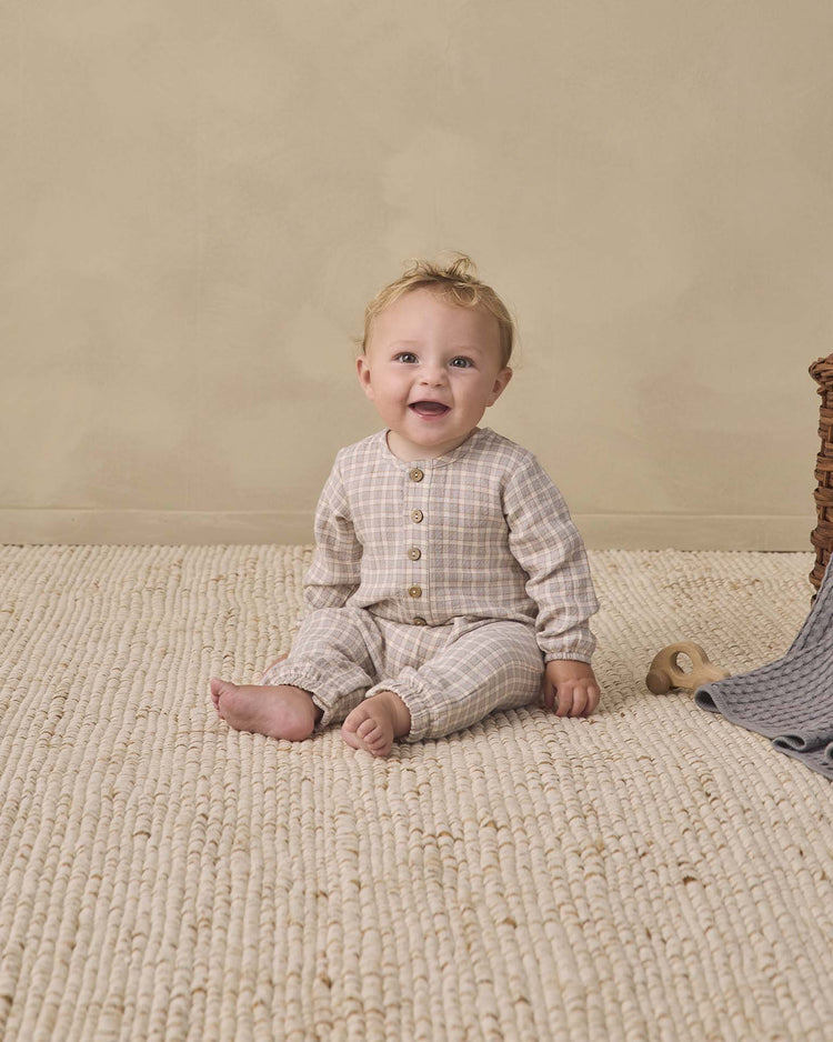 A baby wearing the quincy mae woven jumpsuit in blue gingham sits smiling on a textured cream rug, with a wooden toy and a gray blanket nearby. The background is a plain beige wall.