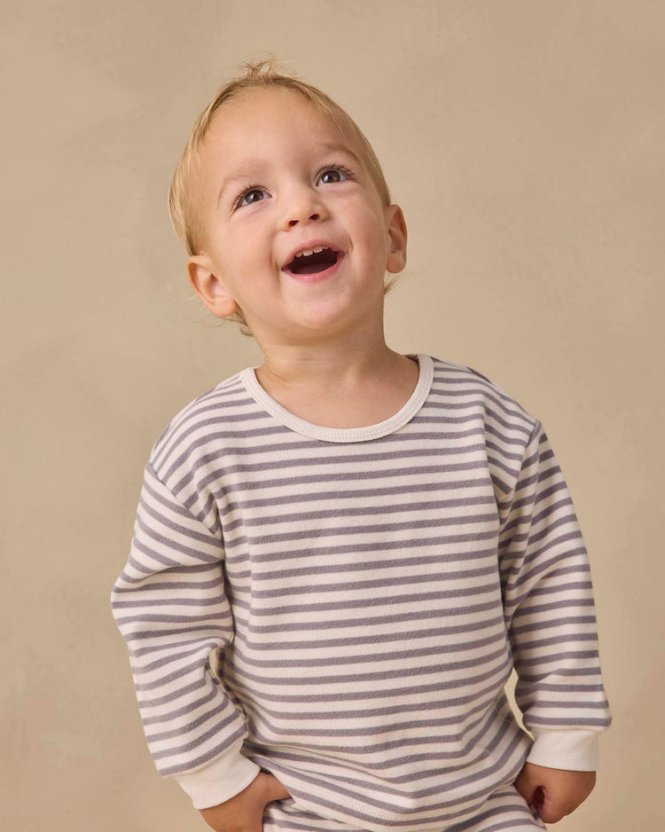 A smiling toddler in the quincy mae cozy set in slate stripe stands against a beige background, looking upward with hands tucked into the drawstring pants. Crafted from soft organic cotton, this outfit offers all-day comfort.
