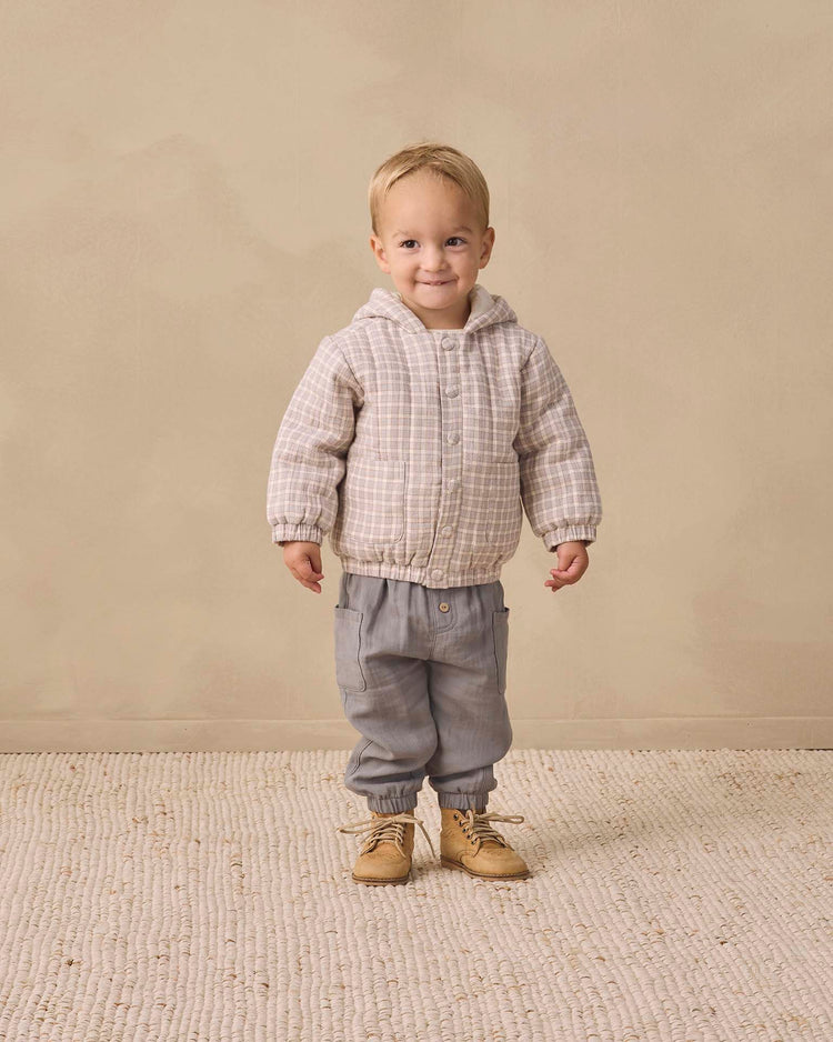 A small child stands smiling, wearing the quincy mae hooded woven jacket in blue gingham made from organic cotton, paired with gray pants with elastic cuffs and tan shoes, on a textured beige rug with a plain beige background.