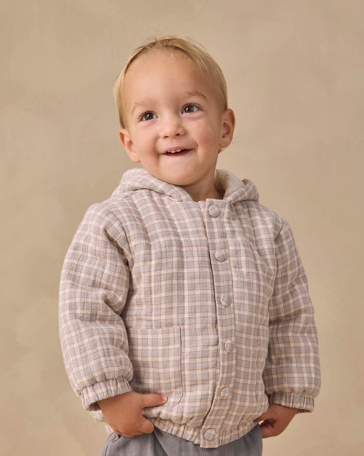 A young child with short blond hair smiles, wearing the quincy mae hooded woven jacket in blue gingham made from organic cotton, hands in pockets, standing against a plain beige background.