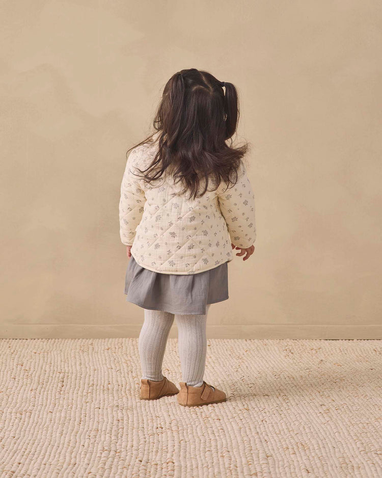 A young child with long dark hair in pigtails stands facing away, wearing a cream jacket, grey skirt, quincy mae tights in cloud, and brown shoes on a textured beige rug with a plain beige wall background.