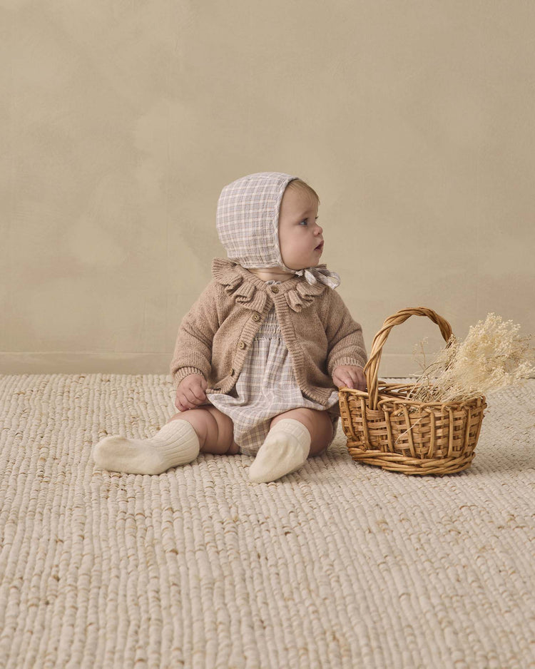 A baby wearing the quincy mae woven baby bonnet in blue gingham, along with a cozy sweater and socks, sits on a textured rug next to a wicker basket filled with dried flowers, gazing to the side against a beige background.