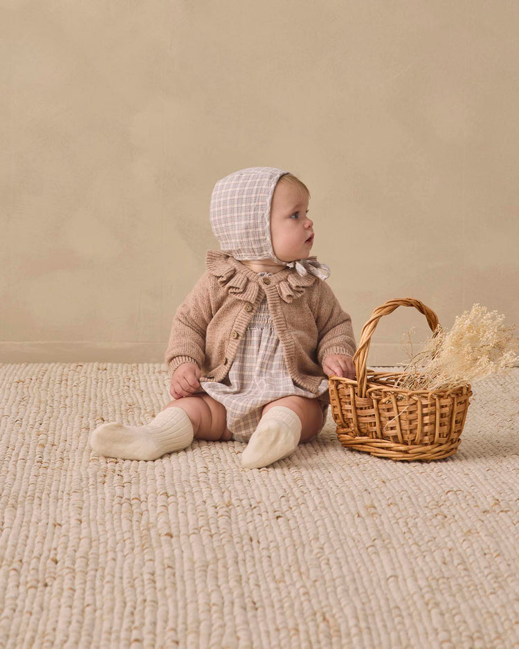 A baby wearing the quincy mae eden romper in blue gingham, along with a matching bonnet and dress, sits on a textured rug beside a wicker basket filled with dried grasses, gazing to the side against a neutral backdrop.