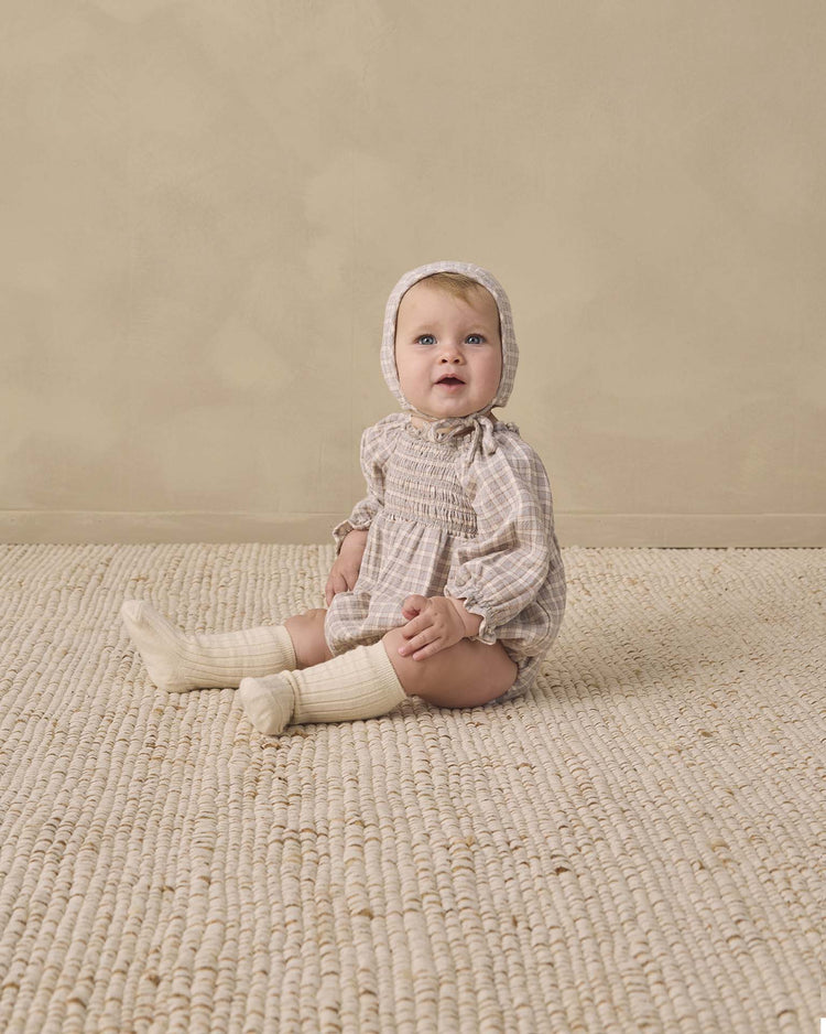 A baby wearing the quincy mae woven baby bonnet in blue gingham sits on a textured cream rug against a beige wall, looking slightly upward.