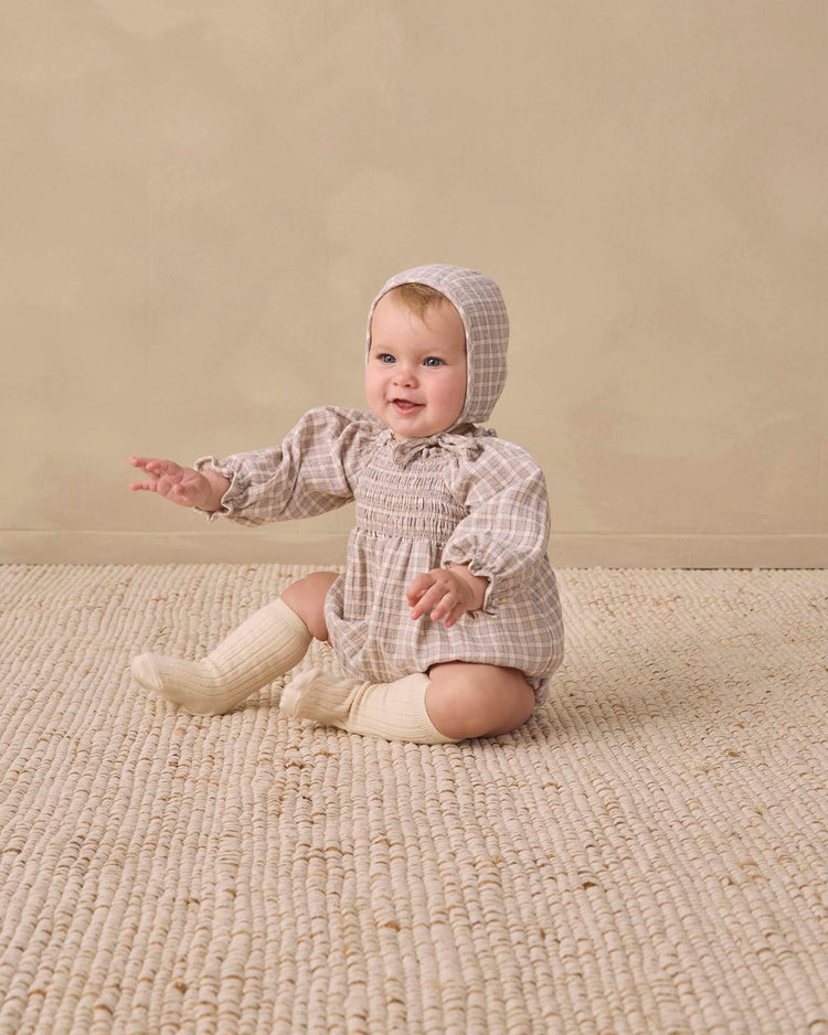 A baby wearing the quincy mae eden romper in blue gingham sits on a textured beige rug against a neutral background, smiling with arms outstretched.