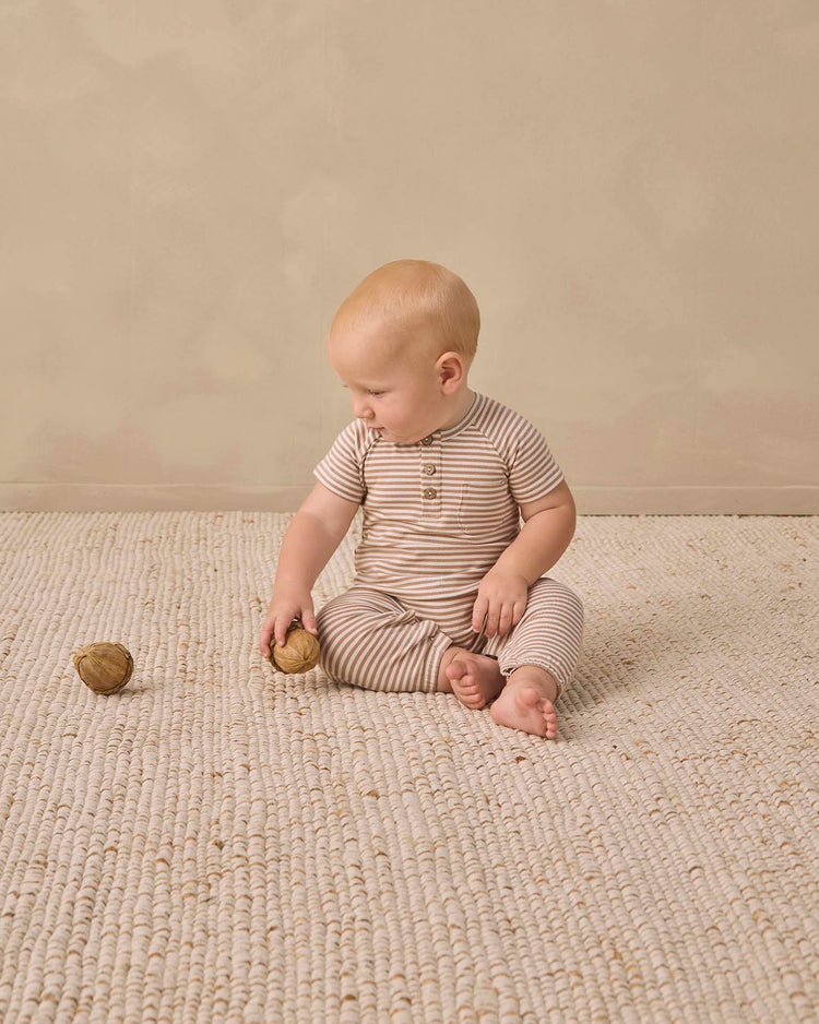 A baby wearing the quincy mae short sleeve jumpsuit in almond stripe sits on a textured cream rug, holding a walnut and gazing at another walnut on the floor. The background is plain beige.
