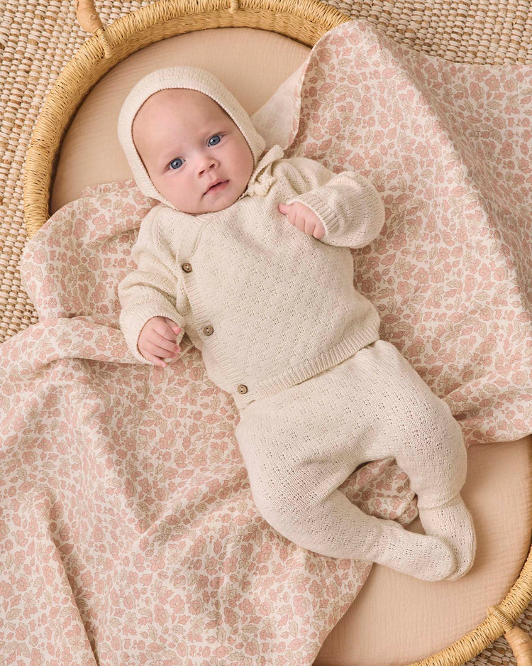 A baby dressed in the quincy mae knit wrap top + pant set in natural, complete with a cozy bonnet, lies on a soft, pink floral-patterned blanket in a woven basket, gazing up with blue eyes.