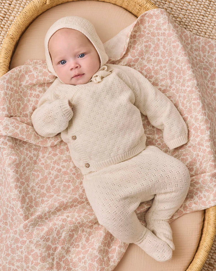A baby in the quincy mae knit wrap top + pant set in natural, crafted from cozy cream organic cotton, lies on a floral-patterned blanket in a woven basket, gazing upward with a calm expression.