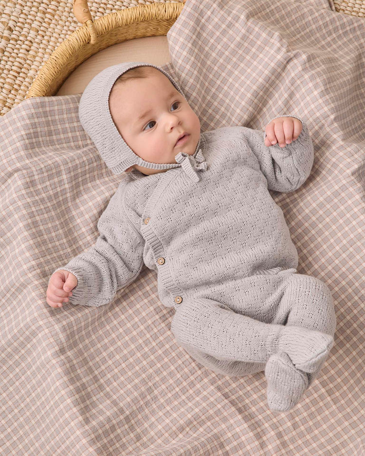 A baby wearing the quincy mae knit wrap top + pant set in cloud and a matching bonnet lies on a beige and plaid-patterned blanket, looking off to the side with one arm raised.