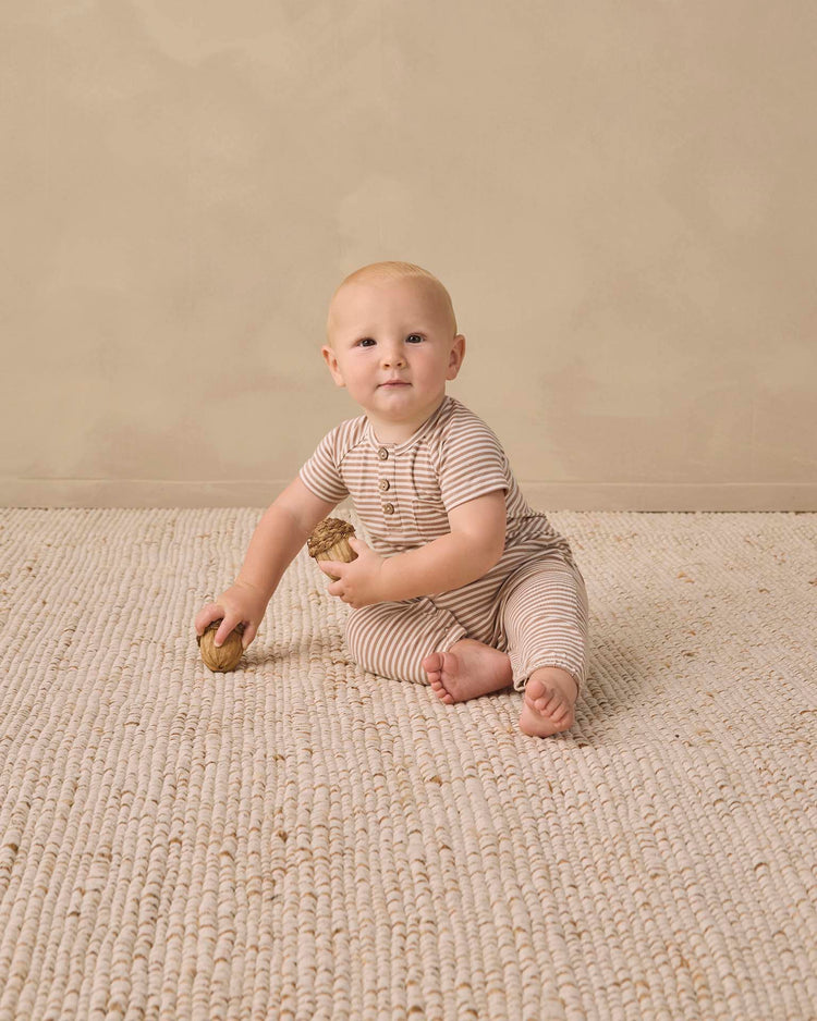 A baby wearing the quincy mae short sleeve jumpsuit in almond stripe sits on a textured beige rug, holding two wooden toys, with a neutral beige wall in the background.