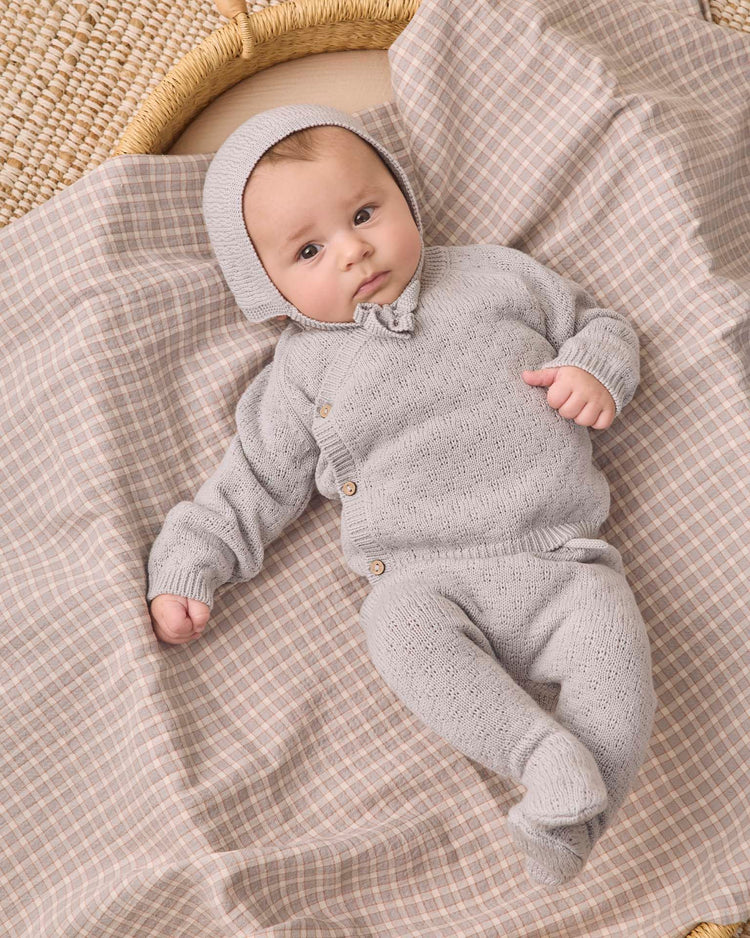 A baby wears the quincy mae knit wrap top + pant set in cloud, complete with a matching bonnet, while lying on a soft, beige plaid blanket beside a woven basket—a cozy scene perfect for showcasing organic cotton baby clothes.