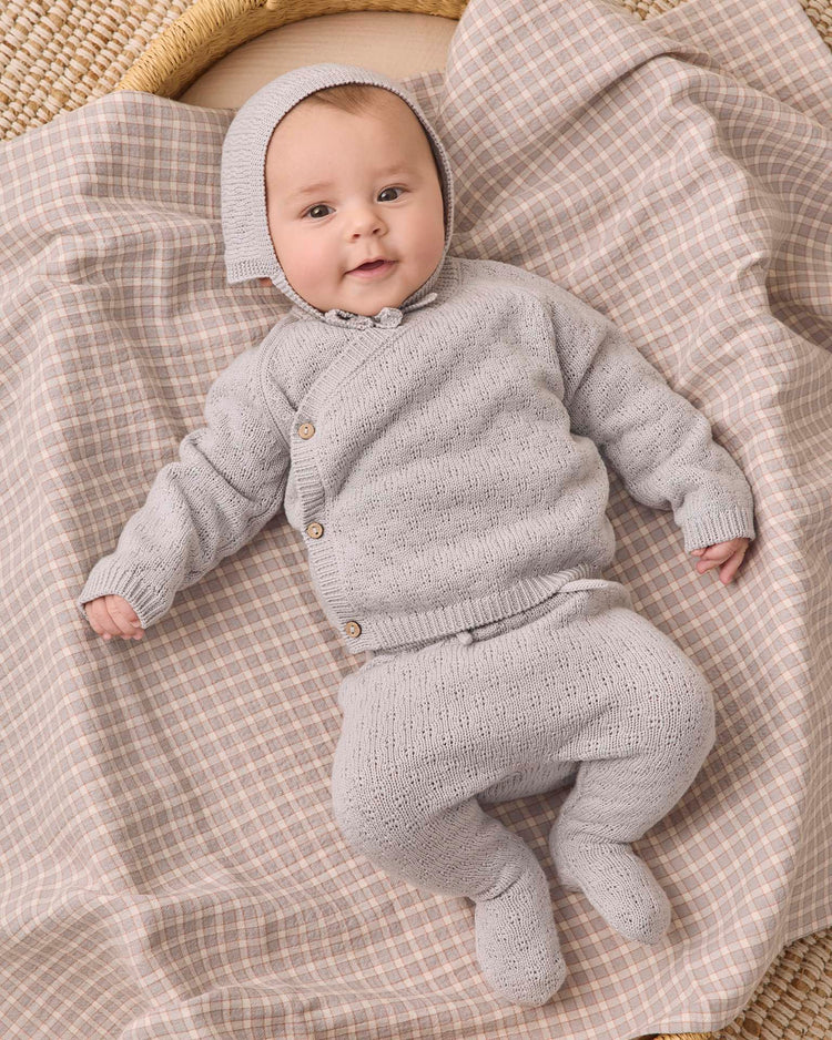 A baby wearing the quincy mae knit wrap top + pant set in cloud and bonnet lies on a beige and gray checkered blanket, looking up with a slight smile. This adorable newborn outfit is crafted from organic cotton for ultimate comfort.