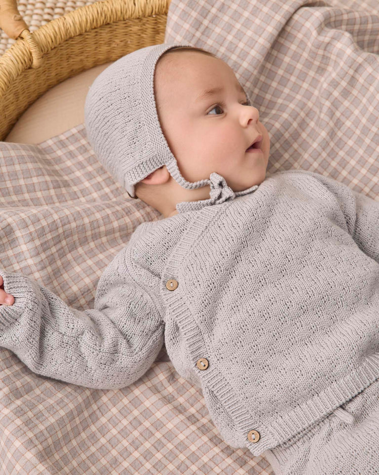 A baby wearing the quincy mae knit bonnet in cloud and a gray sweater lies on a plaid blanket in a wicker basket, gazing to the side with one arm outstretched.