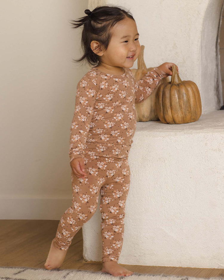 A smiling toddler wearing the quincy mae bamboo pajama set in fall floral stands barefoot indoors, resting a hand on a white surface beside two decorative pumpkins.