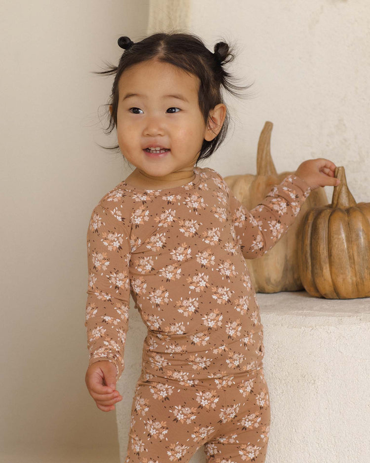 A young child with dark hair styled in small buns wears the Quincy Mae bamboo pajama set in fall floral, smiling and standing near light brown pumpkins on a white surface.