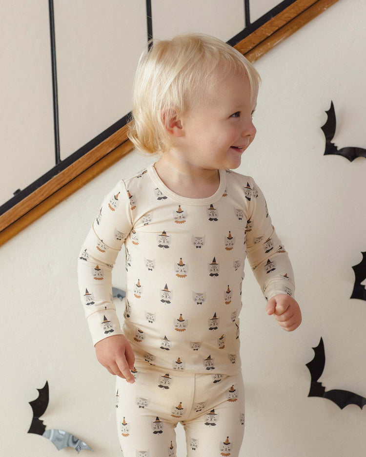 A smiling toddler in the quincy mae bamboo pajama set in costume kittens stands indoors near a staircase decorated with paper bats on the wall, creating a playful Halloween-themed scene.