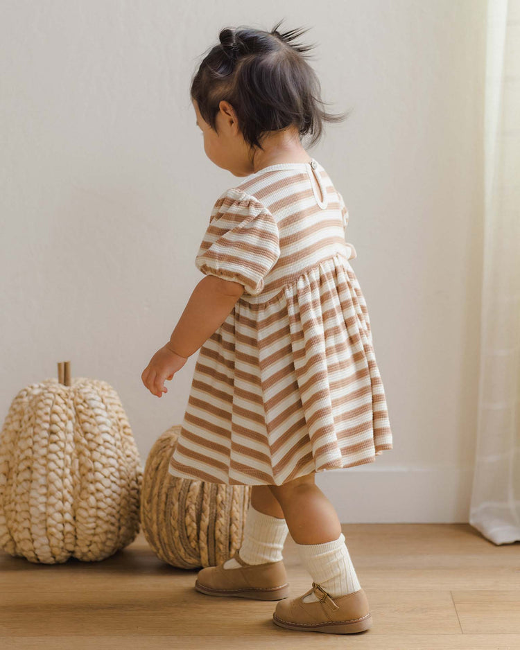 A young child wearing the ‘darla dress in spice stripe’ by quincy mae stands on a wooden floor near two decorative knit pumpkins, facing away from the camera.