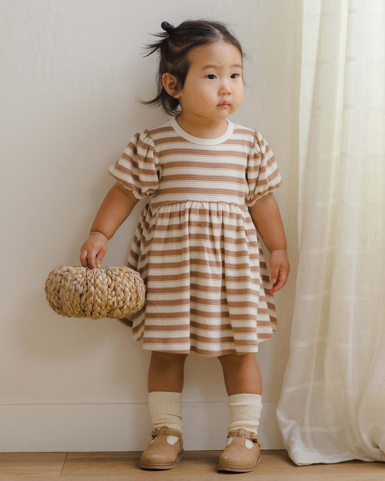 A young child wearing the quincy mae darla dress in spice stripe holds a woven bag while standing by a white curtain. She has dark hair styled in small pigtails, cream socks, and tan shoes, looking slightly to the side.