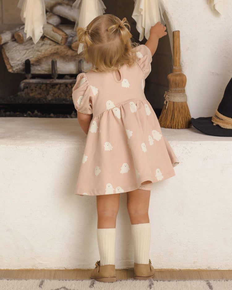 A young girl with blonde hair in pigtails stands on tiptoe, reaching up toward a white mantle, wearing the quincy mae darla dress in sweet ghost—a pink fleece dress adorned with a charming ghost print—with a broom and stacked firewood nearby.