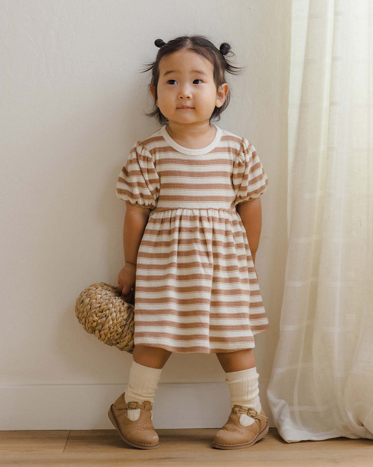 A young child stands indoors near a light curtain, wearing the quincy mae darla dress in spice stripe, paired with tan shoes and cream socks. Her hair is styled in two small buns, and she holds a woven bag.