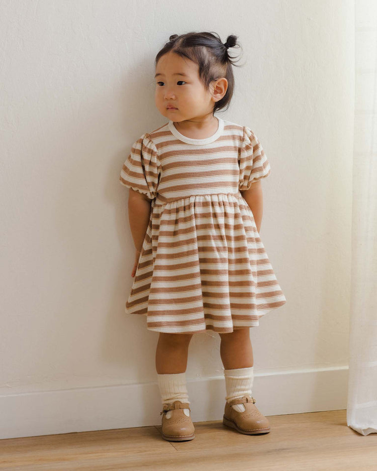 A young child with dark hair in pigtails stands against a light wall, wearing the Quincy Mae Darla Dress in Spice Stripe—an organic cotton dress with puffed sleeves—paired with cream socks and tan shoes, looking slightly to the side.
