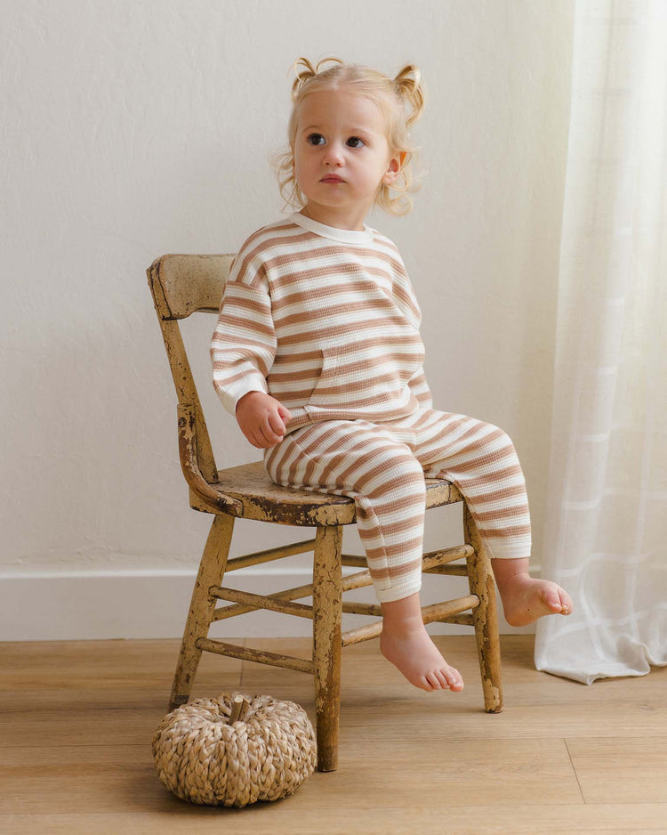 A young child with light hair in pigtails sits barefoot on a rustic wooden chair, wearing the quincy mae waffle slouch set in spice stripe, crafted from soft organic cotton. A woven pumpkin decor rests on the floor nearby as sunlight filters through a sheer-curtained window.