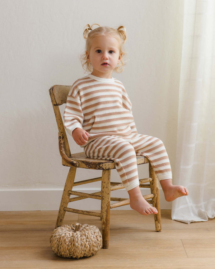 A young child with light hair in pigtails sits barefoot on a worn wooden chair, dressed in the quincy mae waffle slouch set in spice stripe crafted from organic cotton. A woven decorative pumpkin rests on the floor nearby, while gentle light filters through a white curtain.