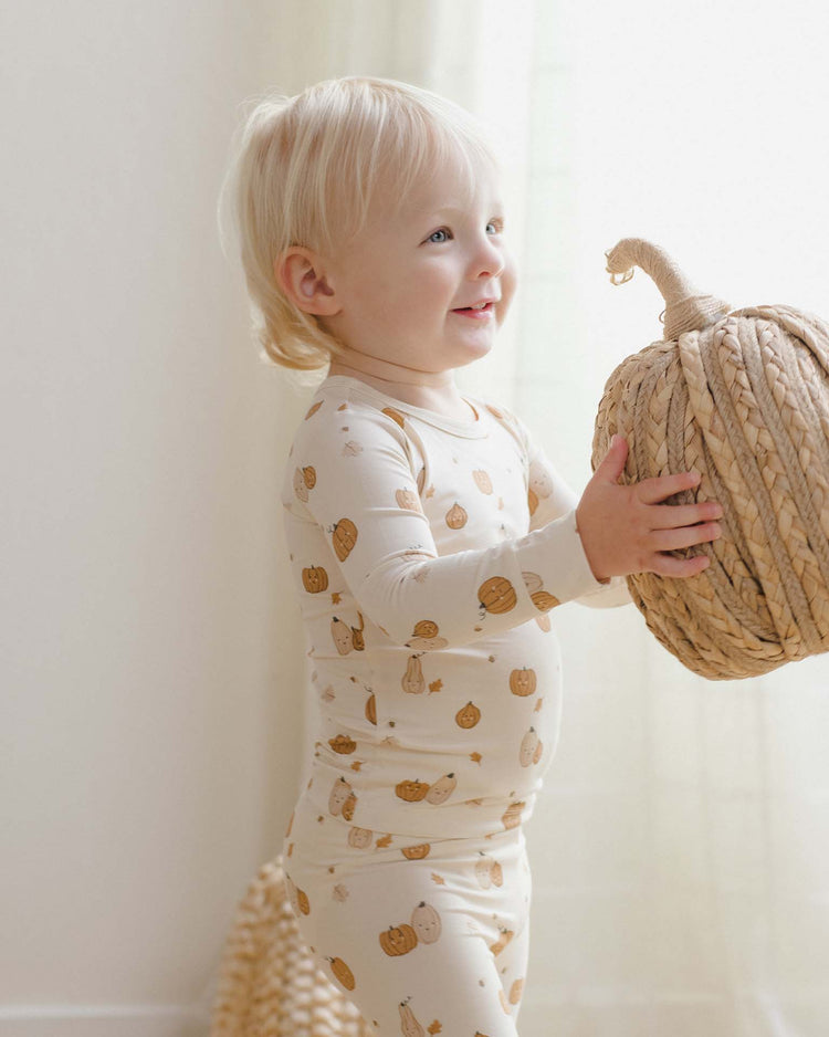 A smiling toddler in a quincy mae bamboo pajama set in harvest friends holds a large woven pumpkin decoration in a softly lit room.