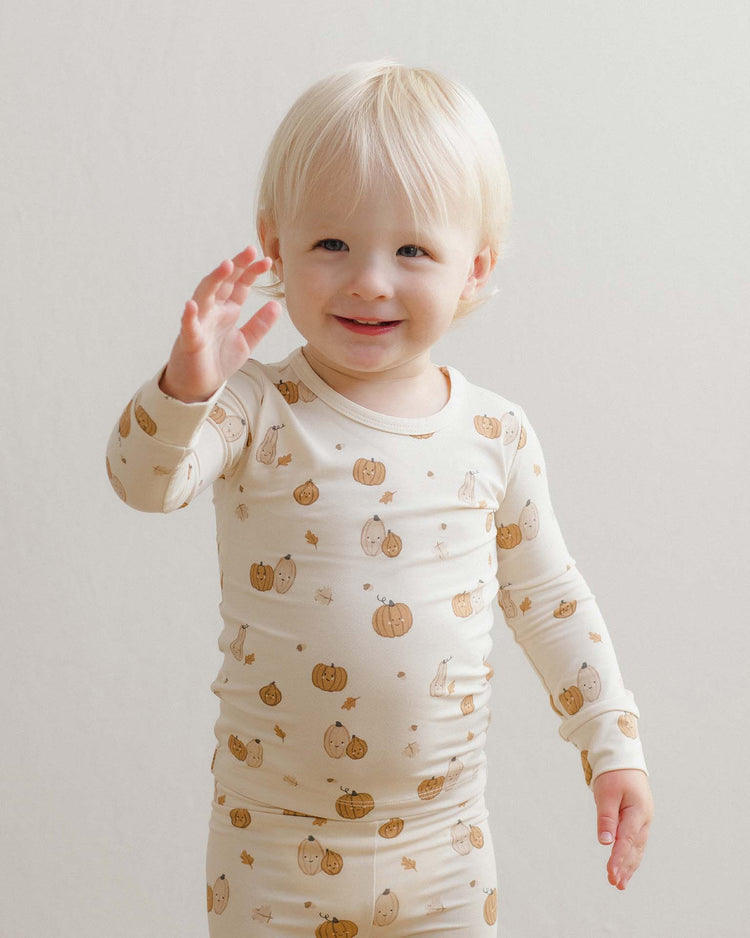A smiling blond toddler wears the quincy mae bamboo pajama set in harvest friends, adorned with playful pumpkin and animal prints, raising one hand as if waving.
