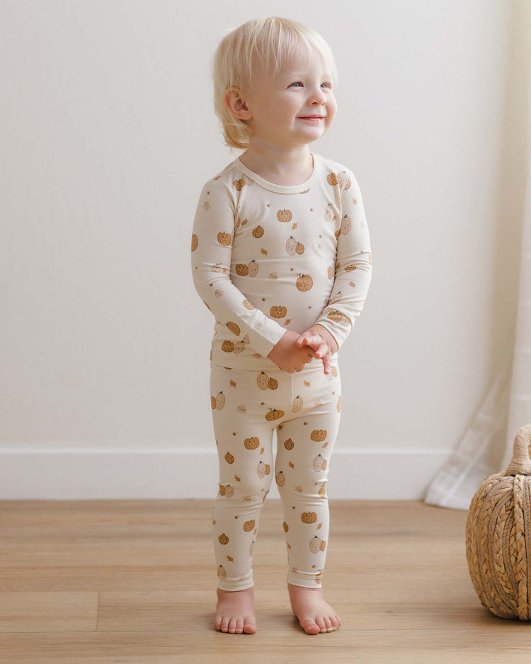 A smiling blond toddler stands barefoot on a wooden floor, wearing the quincy mae bamboo pajama set in harvest friends, surrounded by soft light and with a woven basket nearby.