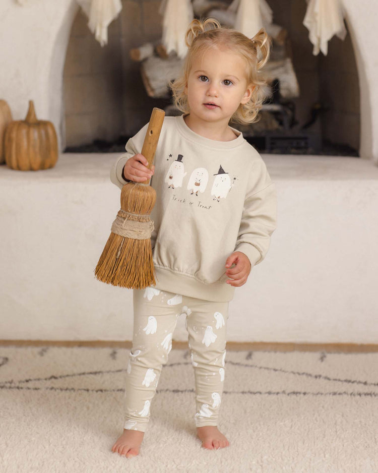 A young child with blonde hair holds a broom, wearing the quincy mae relaxed fleece sweatshirt in trick or treat, featuring playful ghost prints, and stands indoors near a fireplace and decorative pumpkins.