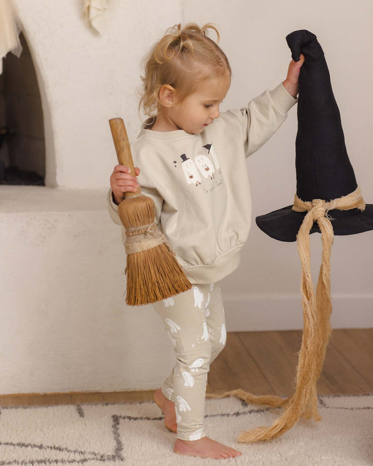 A young child holding a broom in one hand and a black witch hat in the other stands indoors on a rug, dressed for Halloween in the quincy mae relaxed fleece sweatshirt in trick or treat.