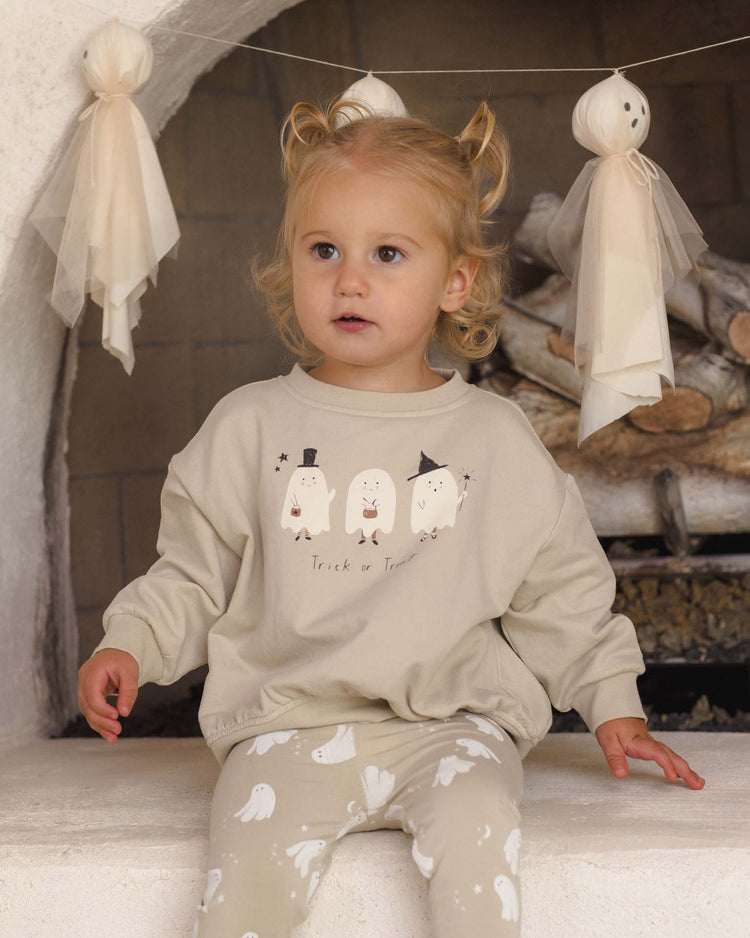 A young child with light hair sits in front of a fireplace, wearing the Quincy Mae relaxed fleece sweatshirt in Trick or Treat. Handmade ghost decorations hang above on a string, and logs are visible in the background.