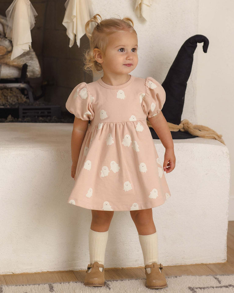 A toddler with blond hair in pigtails wears the Quincy Mae Darla Dress in Sweet Ghost, made from organic cotton, along with cream socks and tan shoes, standing indoors by a white fireplace decorated for Halloween.