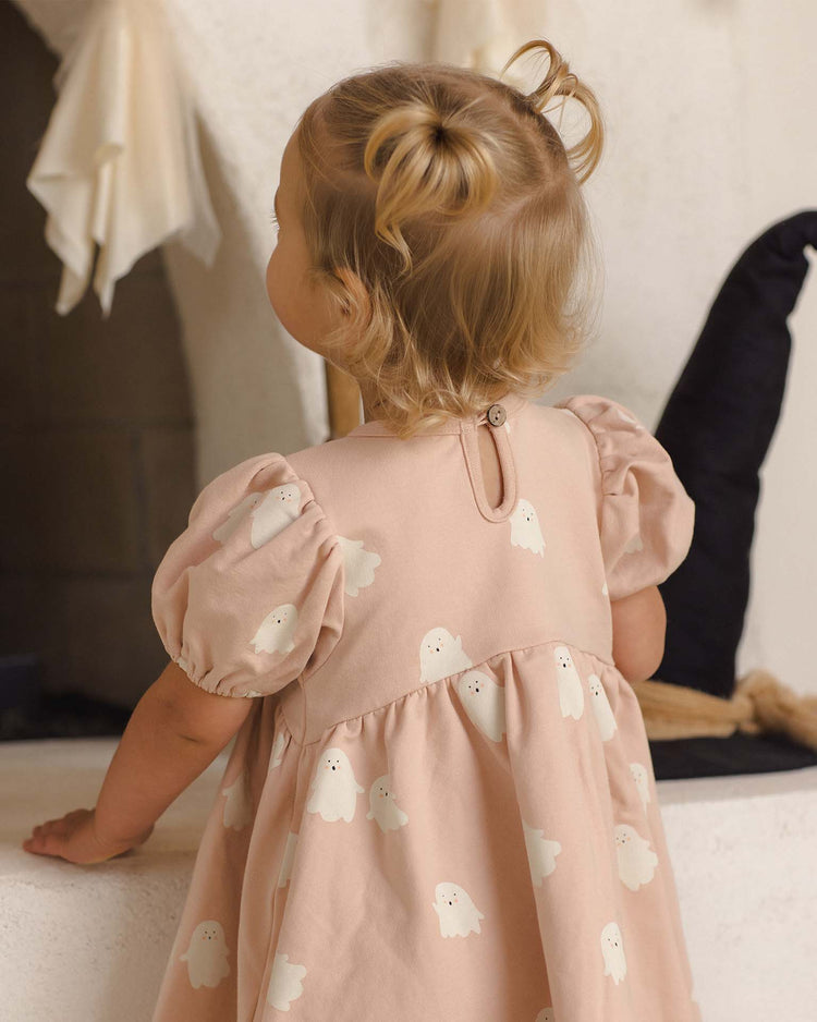 A toddler with light hair in pigtails, wearing the quincy mae darla dress in sweet ghost, stands with her back to the camera, looking toward a white wall and a black witch hat.