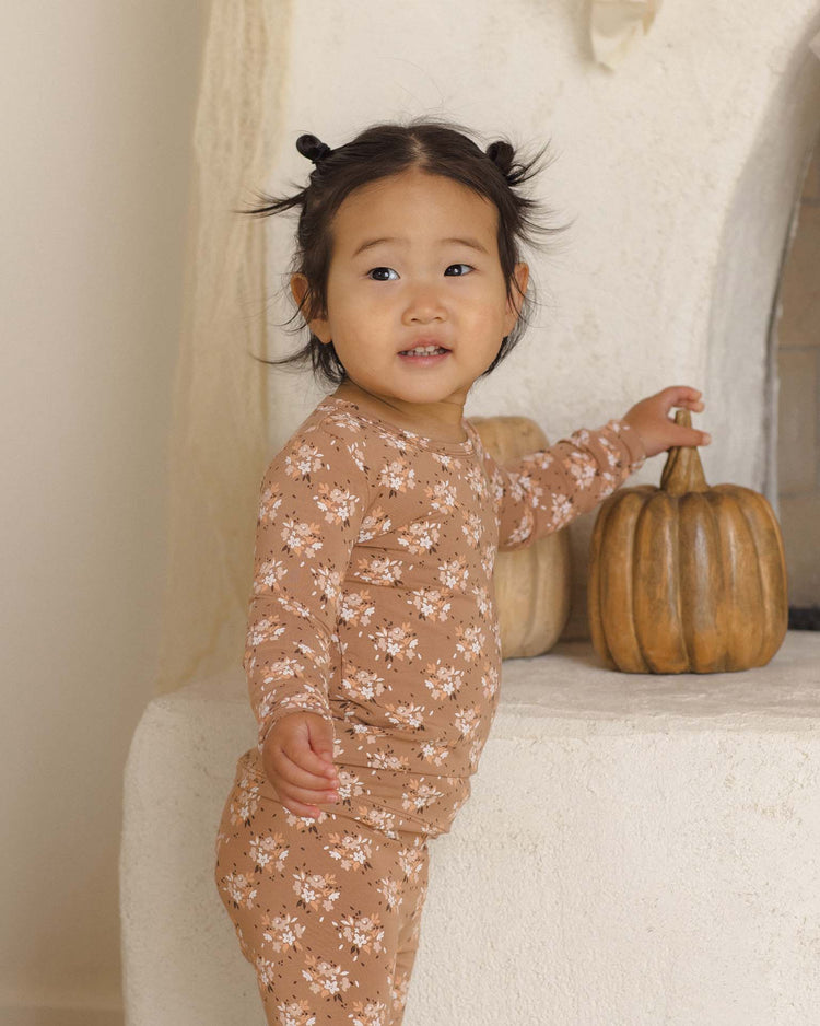 A young child in the Quincy Mae bamboo pajama set in fall floral stands by a white fireplace, gently touching a decorative pumpkin and smiling softly.