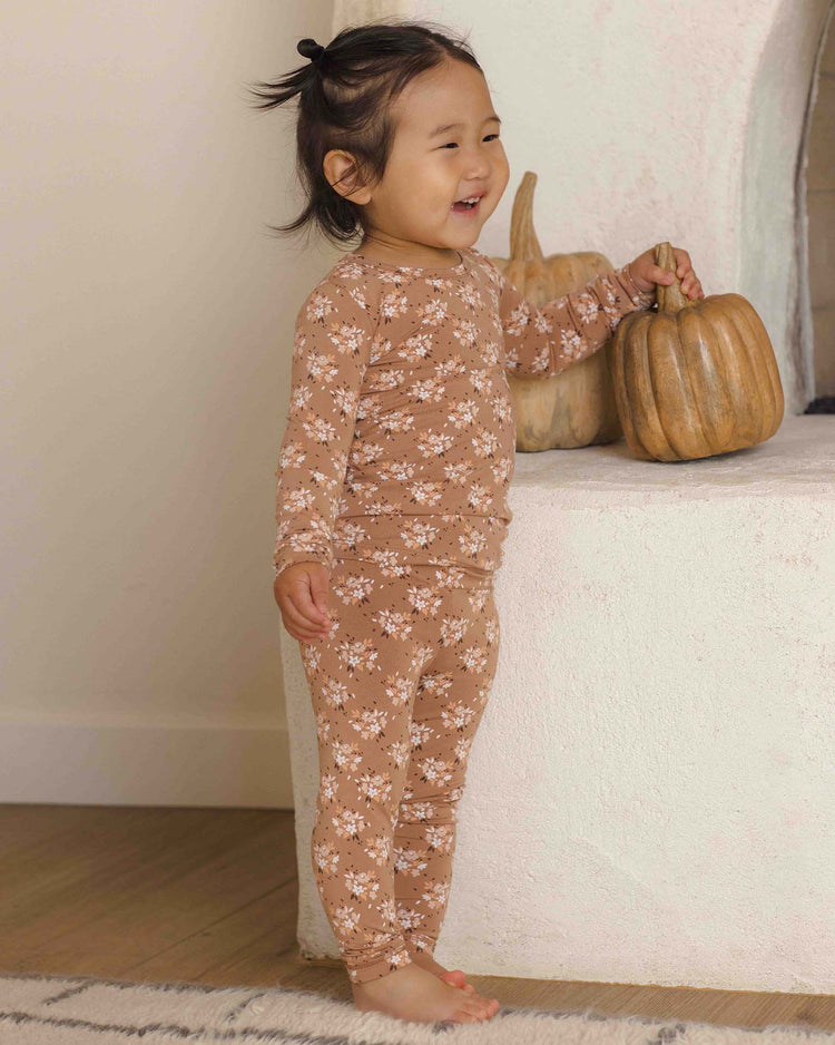 A smiling toddler dressed in the quincy mae bamboo pajama set in fall floral stands barefoot beside a white fireplace, gently touching a decorative pumpkin.