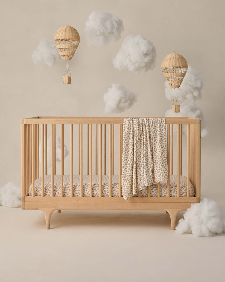 A wooden baby crib draped with the quincy mae bamboo swaddle in wildflower stands against a beige backdrop, surrounded by soft artificial clouds and two wicker hot air balloon decorations hanging above.