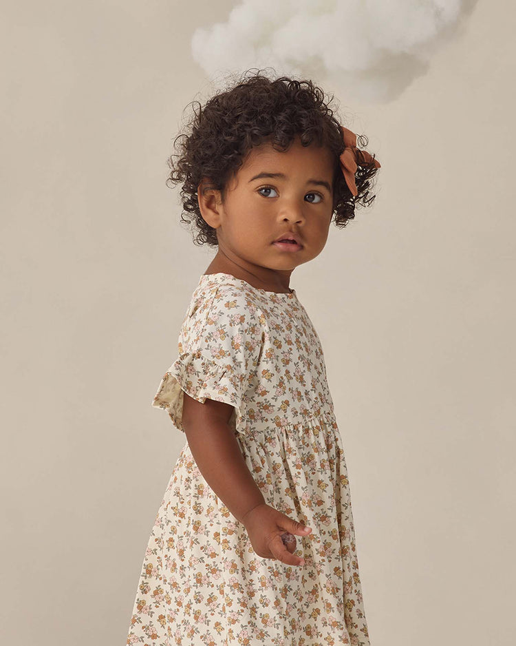 A young child with curly hair wears the quincy mae bamboo babydoll dress in wildflower, standing before a beige background and looking at the camera, with a small white cloud suspended above their head.