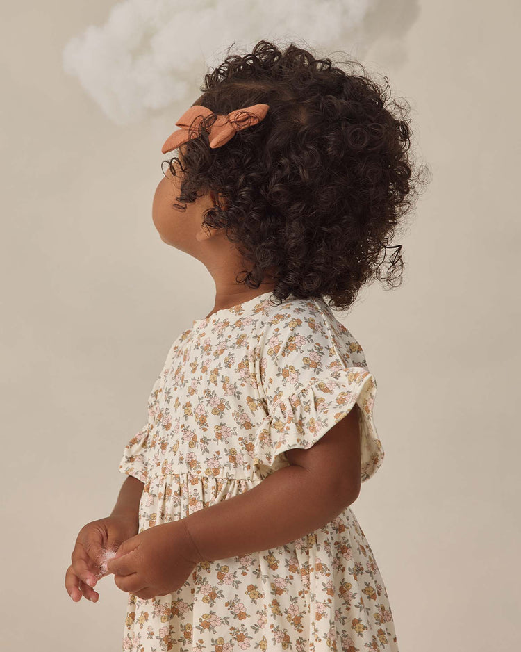 A young child with curly hair and a peach bow looks to the side, wearing the quincy mae bamboo babydoll dress in wildflower. The cream dress features a floral pattern and ruffle sleeves, set against a neutral background with a small cloud above her head.