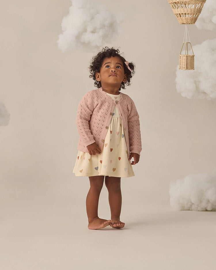 A young child with curly hair stands barefoot on a light floor wearing the quincy mae darla dress in confetti hearts, paired with a pink waffle knit cardigan. Fluffy clouds and a small basket decorate the soft, neutral-toned background.